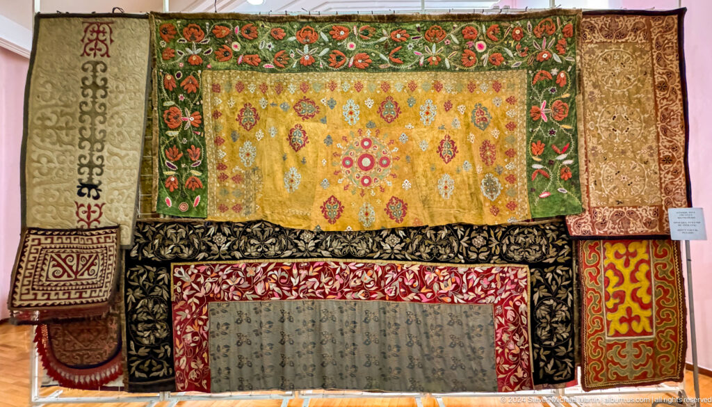Medium view of colorful Central Asian carpets and embroidered textiles mounted on a red display wall in the Central State Museum in Almaty, Kazakhstan.
