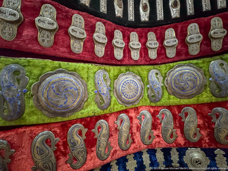 Close-up of red and green textile belts covered with metal plaques against a red display backing in the Central State Museum of Kazakhstan in Almaty.