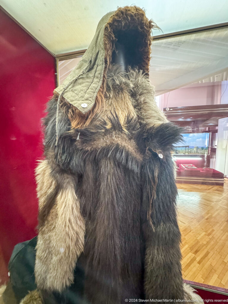 Medium view of a traditional brown fur coat with decorative trim displayed against a red background at the Central State Museum of Kazakhstan in Almaty.