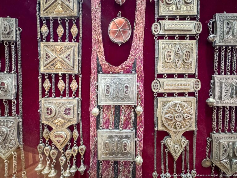 Museum display case with vertically arranged silver jewelry and pendants against a neutral background.