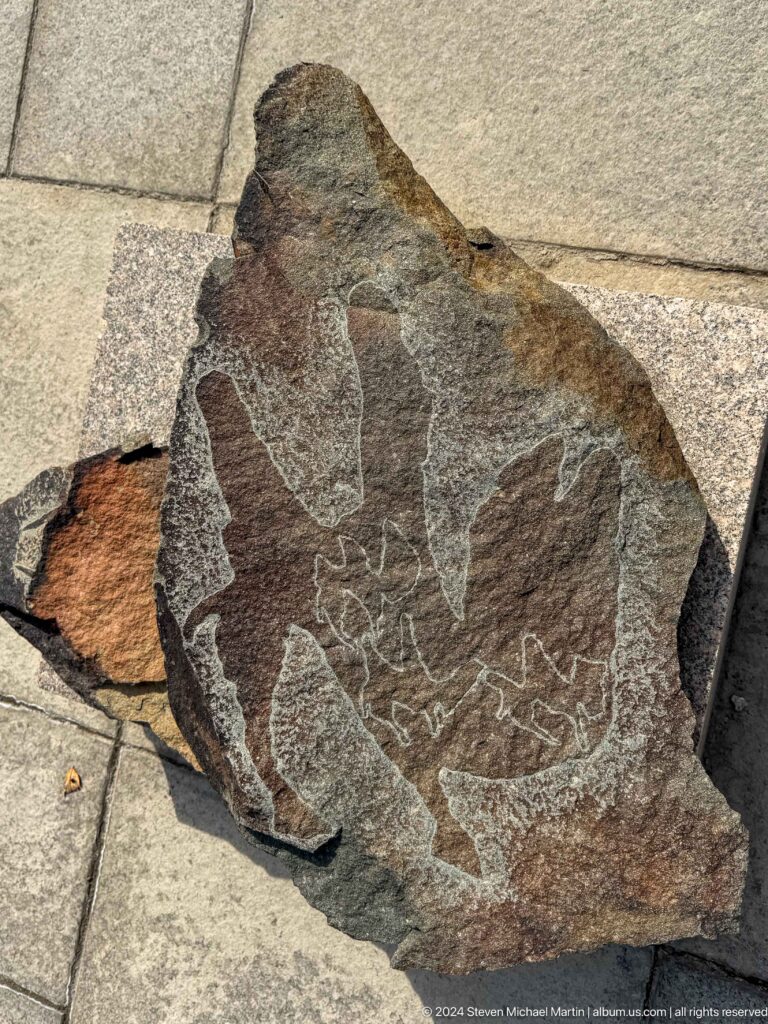 Medium view of a dark, broken stone fragment with faint carved lines on its rough surface.
