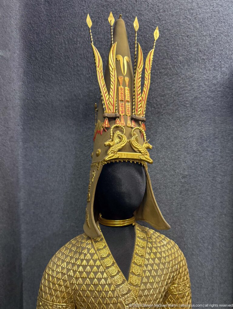 Medium view of a tall, pointed ornate ceremonial helmet with prominent gold-colored decoration on display in a museum.