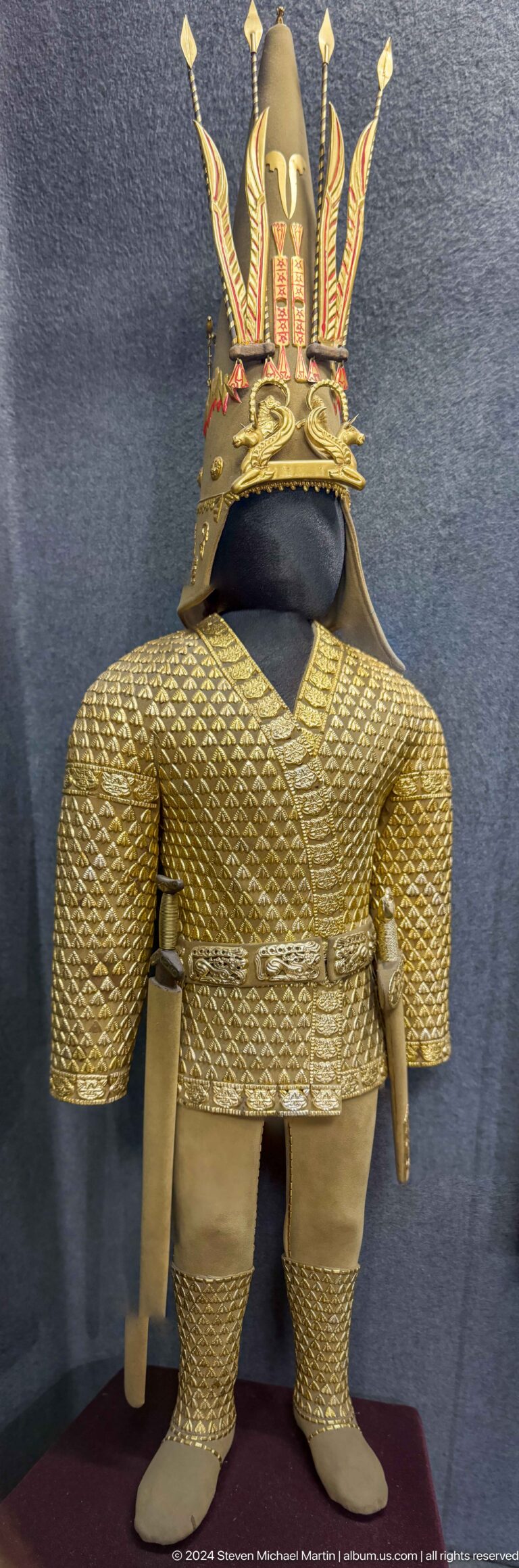 Close-up of a gold-colored scale-patterned armored tunic against a red museum display backing in the Central State Museum of Kazakhstan in Almaty.