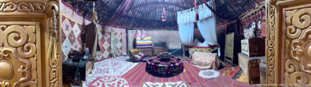 Interior of a Kazakh yurt in a museum, with carpets on the floor, patterned textiles on the walls, and a low table at the center.