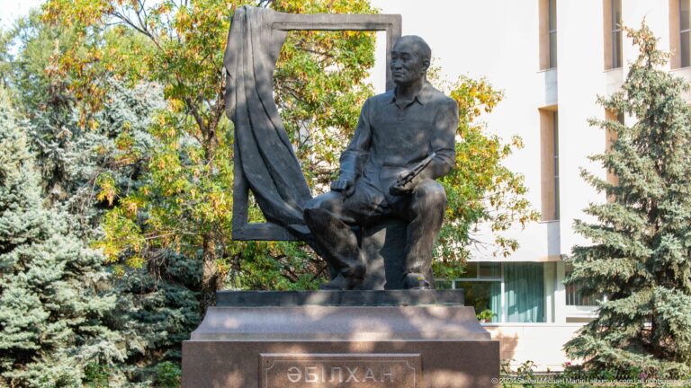 Dark seated male figure on outdoor pedestal.