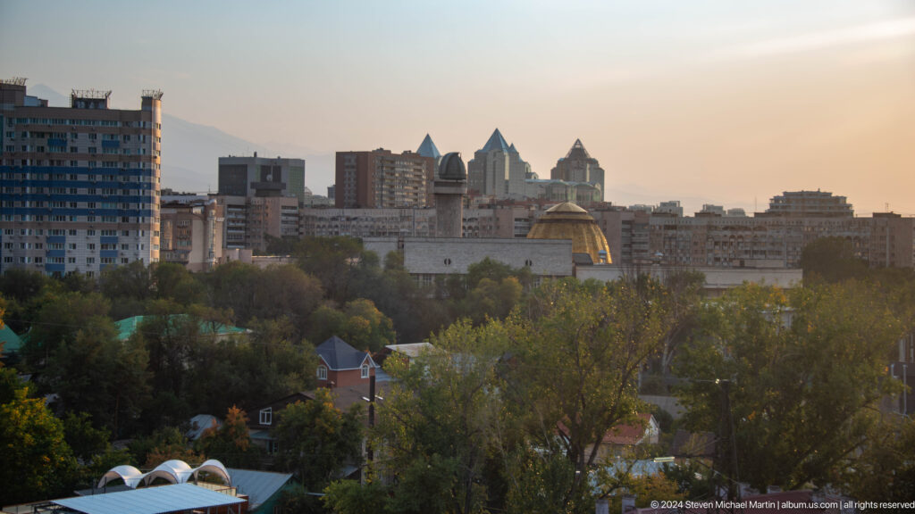 Telephoto view of central Almaty’s skyline with distinctive peaked towers seen from Kok Tobe above a wooded foreground.