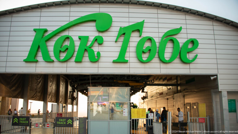 Main public entrance to the Kok Tobe hilltop recreation complex with green Kok Tobe sign above turnstiles and visitors.