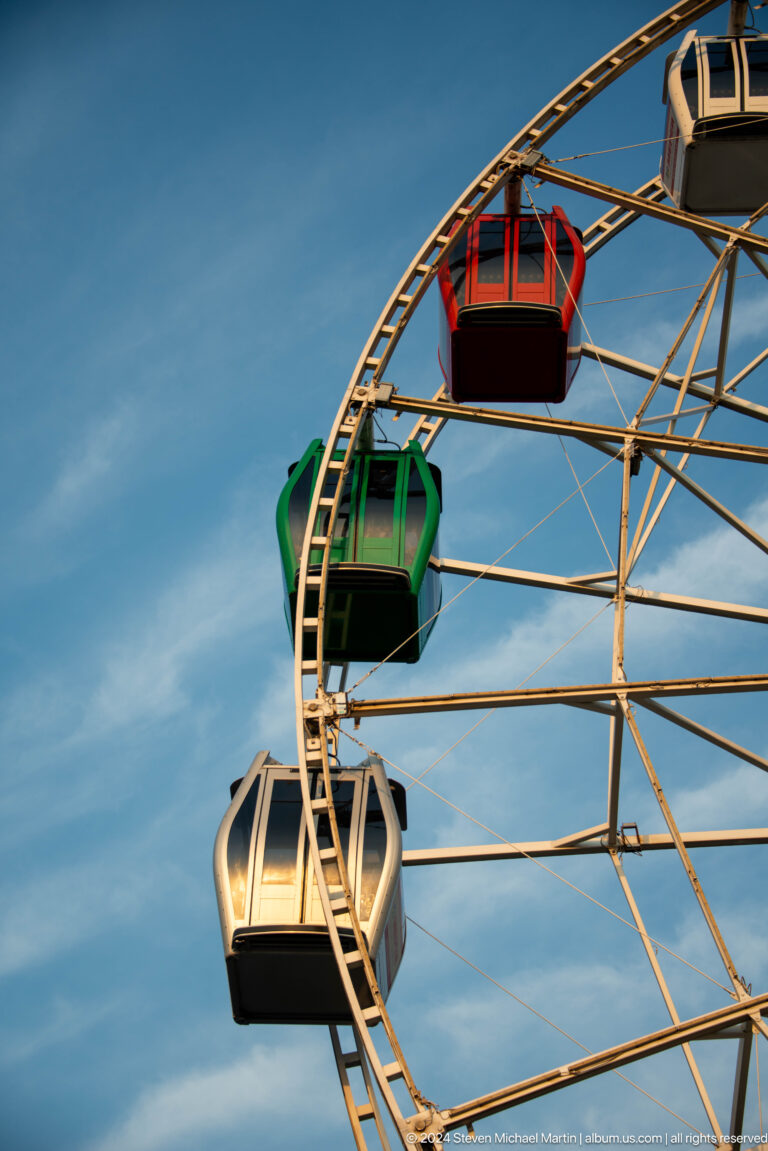 Enclosed green Ferris wheel cabin hanging within the white metal spokes and frame of the Kok Tobe wheel against a blue sky.
