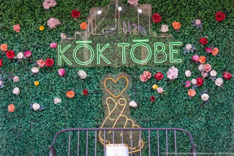 Floral selfie wall at Kok Tobe with neon ‘KOK TOBE’ sign and illuminated hand-heart symbol surrounded by artificial flowers.