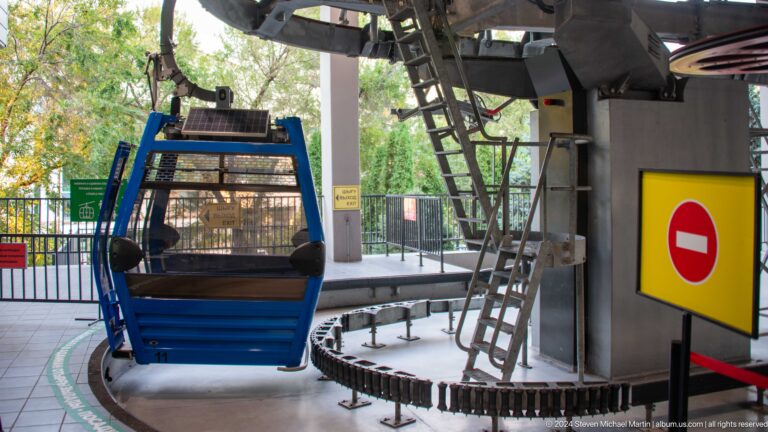 Blue enclosed Kok Tobe cable-car gondola on the loading track inside the station, with surrounding machinery and guide rails.