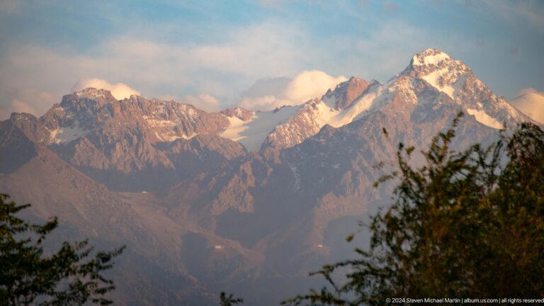 Telephoto view of rugged, snow-streaked Tian Shan peaks in late-afternoon light, seen from Kok Tobe above Almaty.