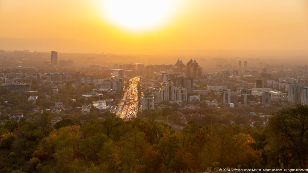 Elevated sunset view from Kok Tobe over central Almaty with a glowing main avenue and tree-covered hill in the foreground.