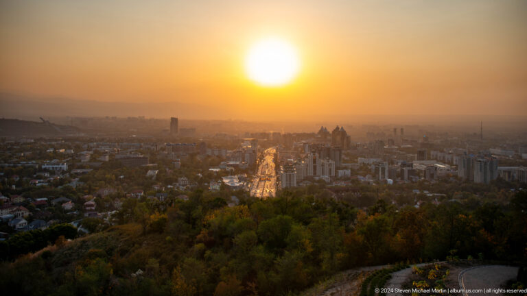 Wide sunset panorama over Almaty from the Kok Tobe viewpoint, with the sun low in an orange sky above the city.