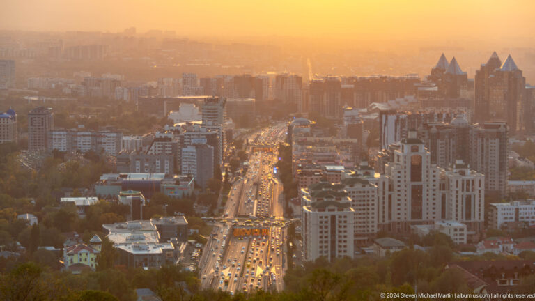 Telephoto view from Kok Tobe of a major Almaty avenue at golden hour, with heavy traffic and tall buildings on both sides.
