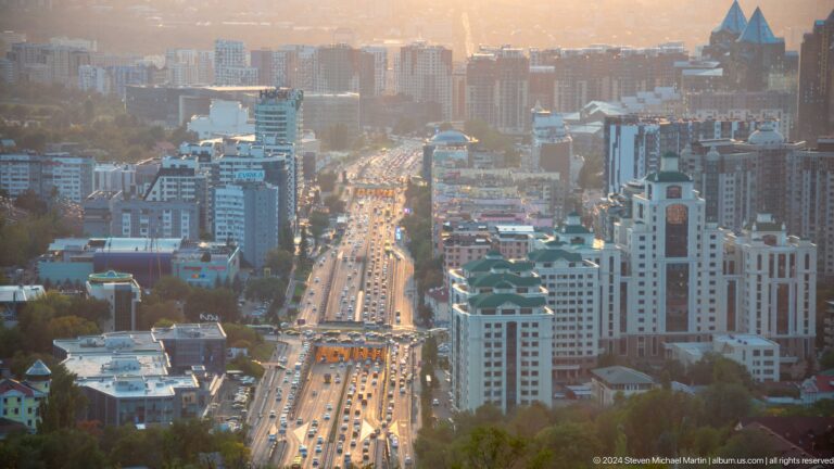 Telephoto view of a central Almaty boulevard lined with high-rise buildings and heavy traffic.