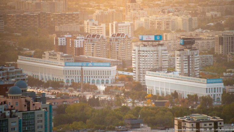 Telephoto view from Kok Tobe of central Almaty, showing a long pale government building in front of dense mid-rise commercial blocks.
