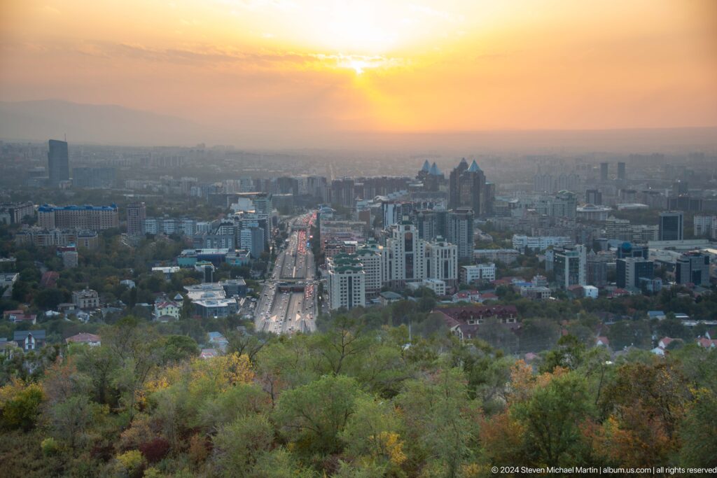 Elevated view of Almaty’s skyline at sunset, with sunlight breaking through clouds and a central avenue glowing through atmospheric haze.