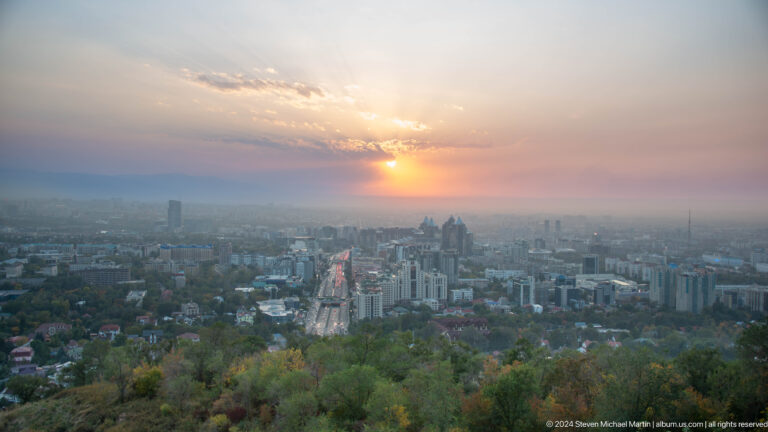 Sunset over Almaty seen from Kok Tobe, with the low sun glowing through clouds above the hazy city below.