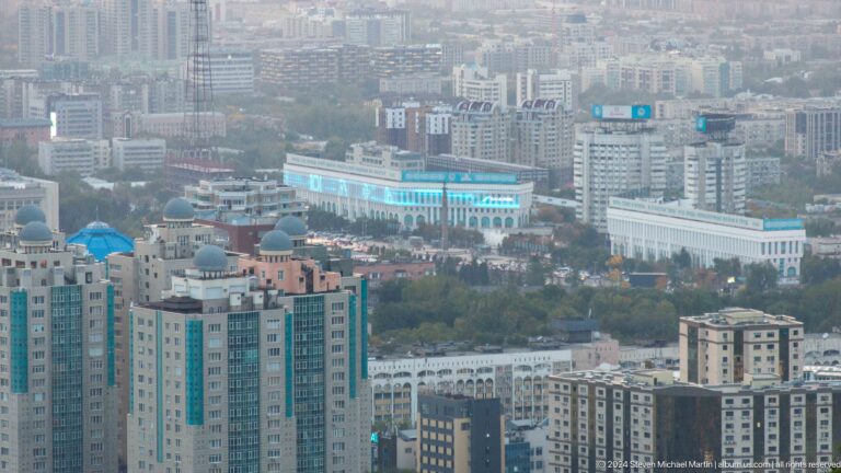 Telephoto view of central Almaty showing a domed residential tower in front of large white civic buildings.