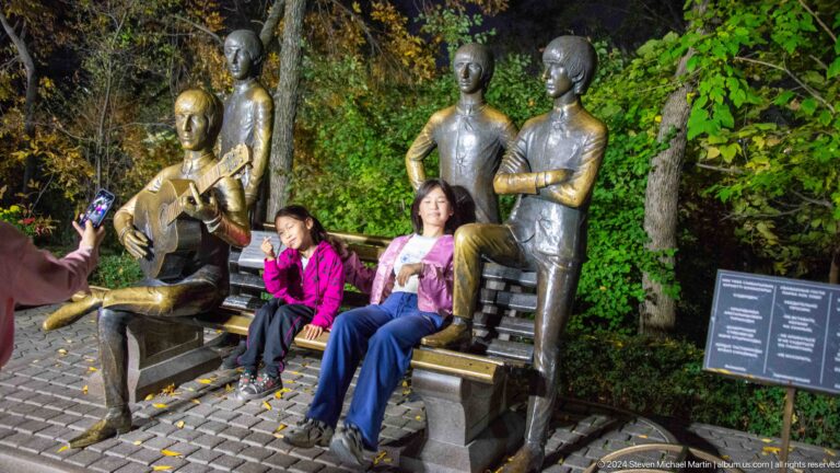 Two visitors seated on a bench among life-size bronze Beatles figures at the Kok Tobe monument.