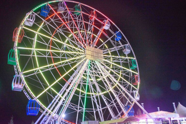Front view of the Kok Tobe Ferris wheel at night, outlined by multicolored LED lights with visible radial spokes and cabins.