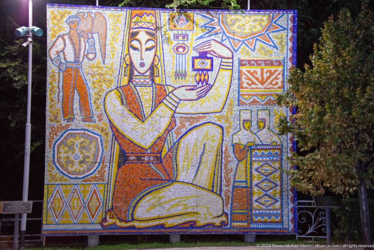 Colorful mosaic mural of a stylized seated Central Asian figure with surrounding sun, vessels, and geometric motifs at Kok Tobe.