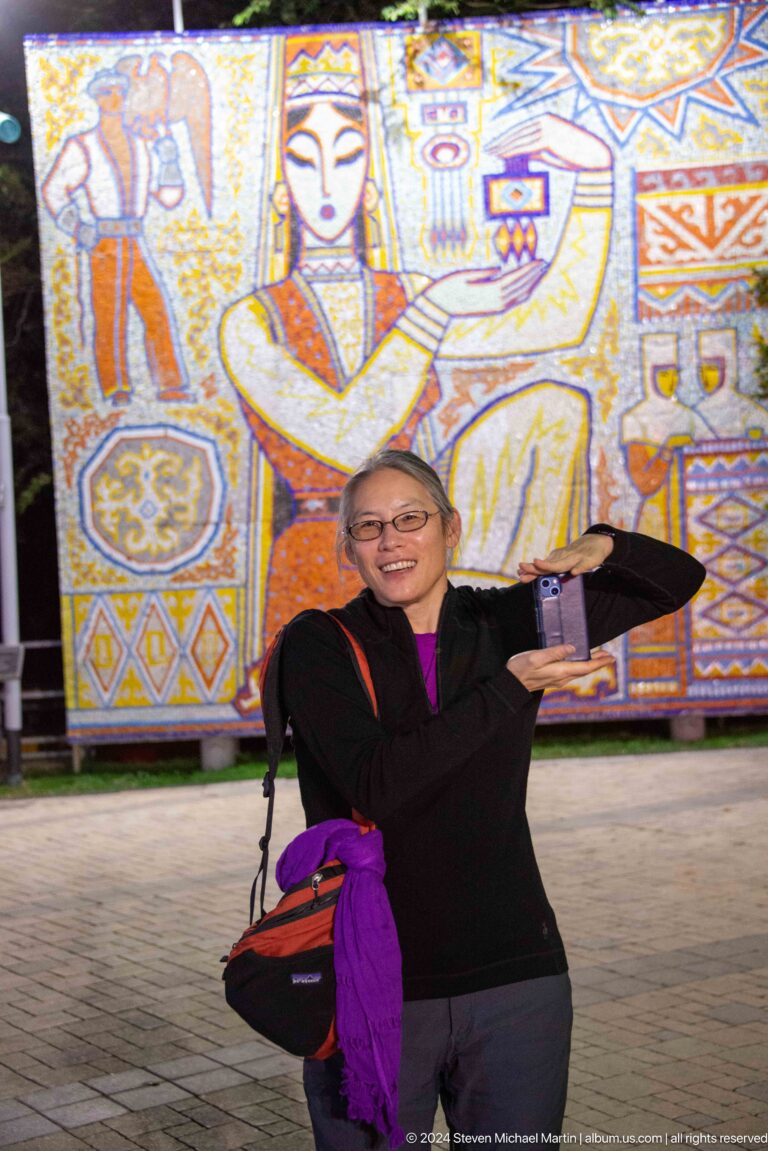Smiling woman in glasses and dark clothing holding up a smartphone as she poses before a decorative mural at Kok Tobe.