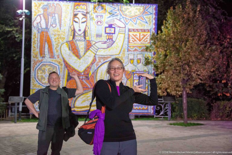 Two visitors pose at night in front of an illuminated Kok Tobe mural.
