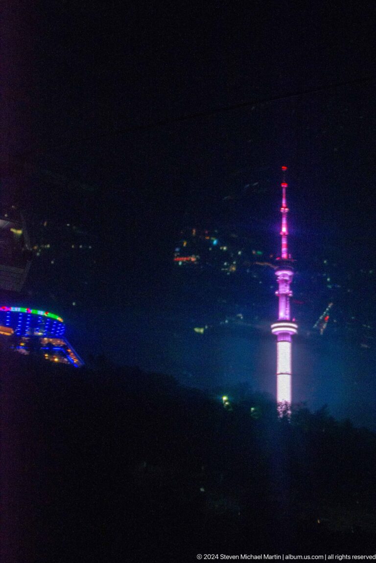 Almaty Television Tower lit pink and white at night, rising above hill lights as seen from Kok Tobe.