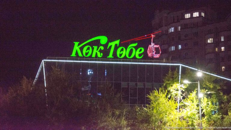 Night view of the Kok Tobe station building with a glowing green Kok Tobe sign and red gondola icon outlined by white lights.