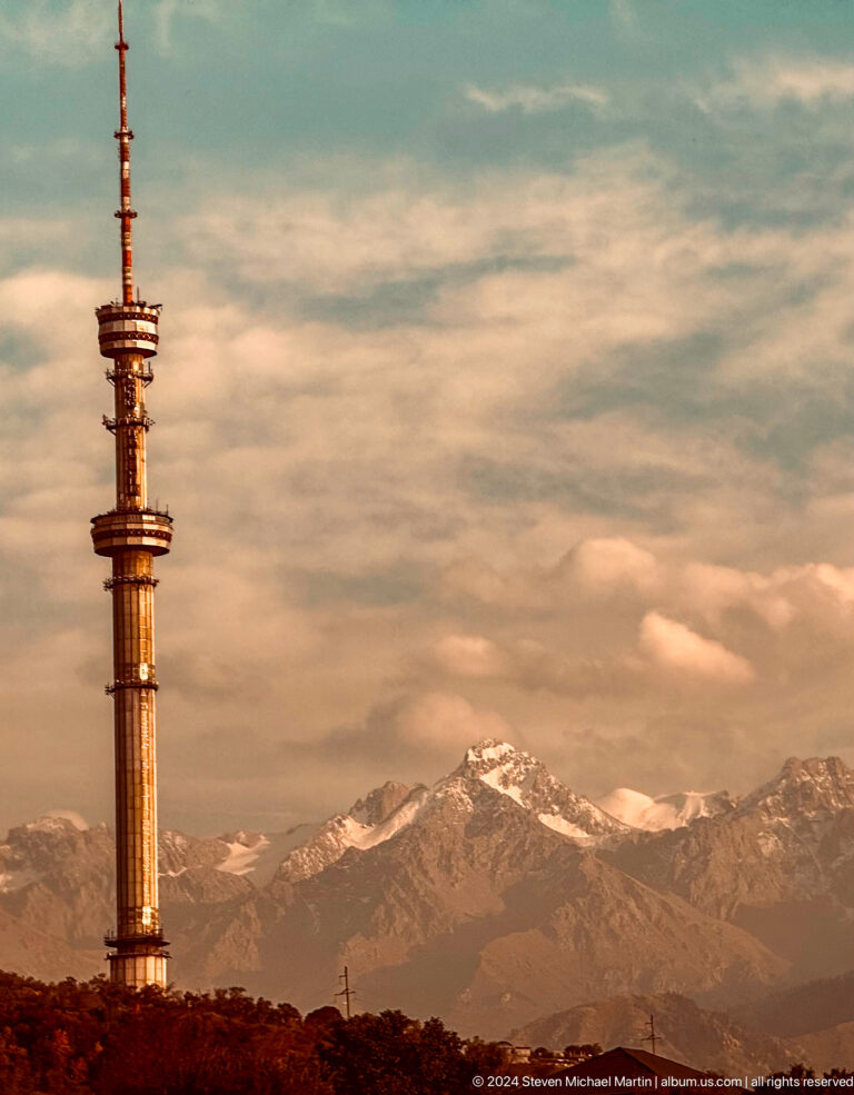 Almaty Television Tower in evening light with a snow-covered mountain peak in the background.
