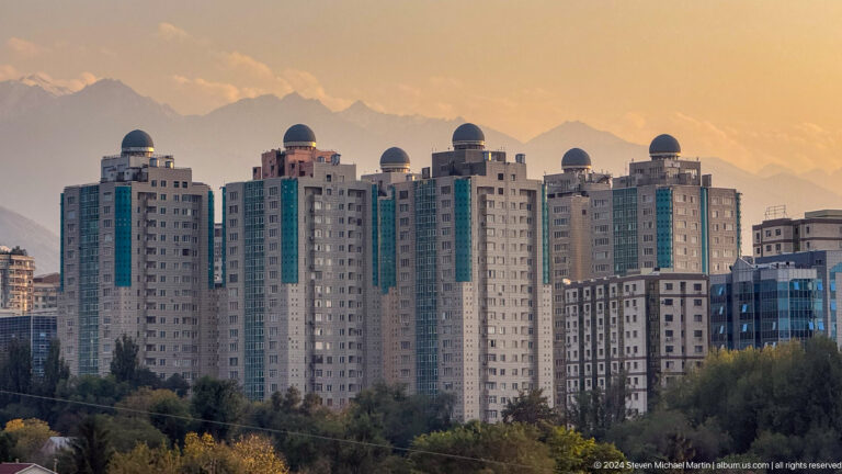 Telephoto view of pale, domed-roof residential towers in Almaty set before hazy mountains.