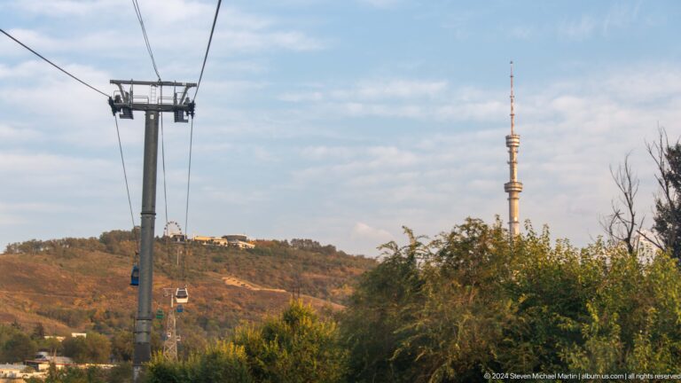 Almaty Television Tower on Kok Tobe hill with cable-car cabins crossing the wooded slope in the foreground.