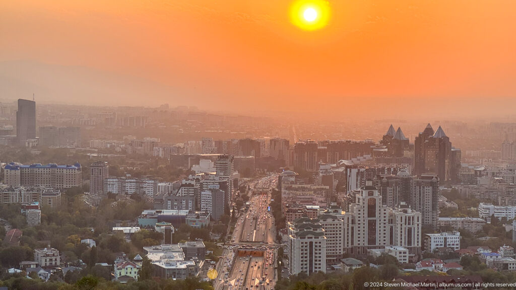 Orange sun setting over central Almaty with a major boulevard leading through dense city development, viewed from Kok Tobe.