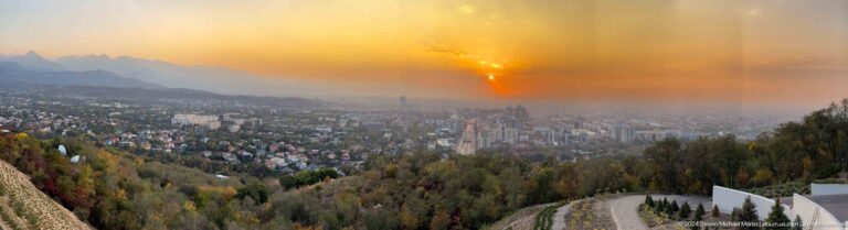 Cloud-filtered sunset over Almaty from the elevated Kok Tobe viewpoint, with hazy distant buildings beneath an orange sky.