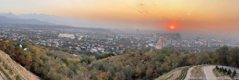 Late red sunset over Almaty from Kok Tobe, with a glowing sun near the horizon above residential districts and central towers.