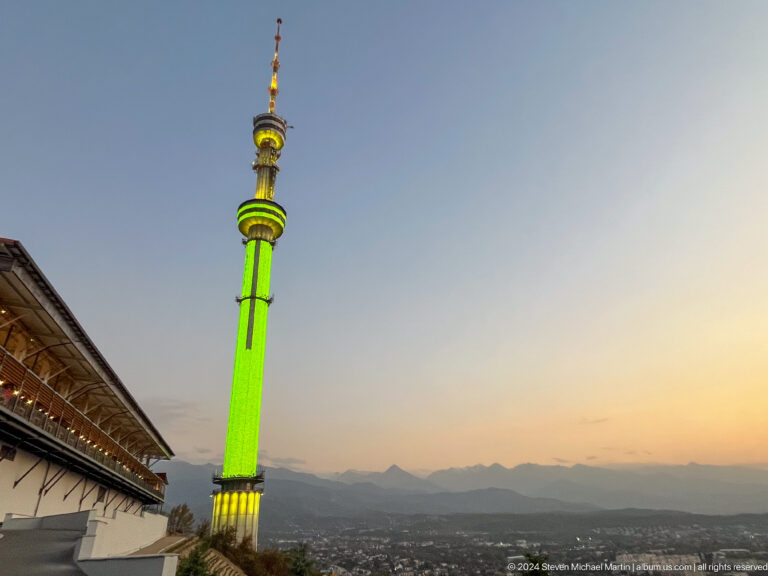 Almaty Television Tower lit green at dusk with mountains visible along the horizon.