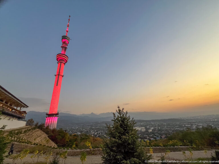 Almaty Television Tower lit in red at sunset above the hillside with mountains in the background.