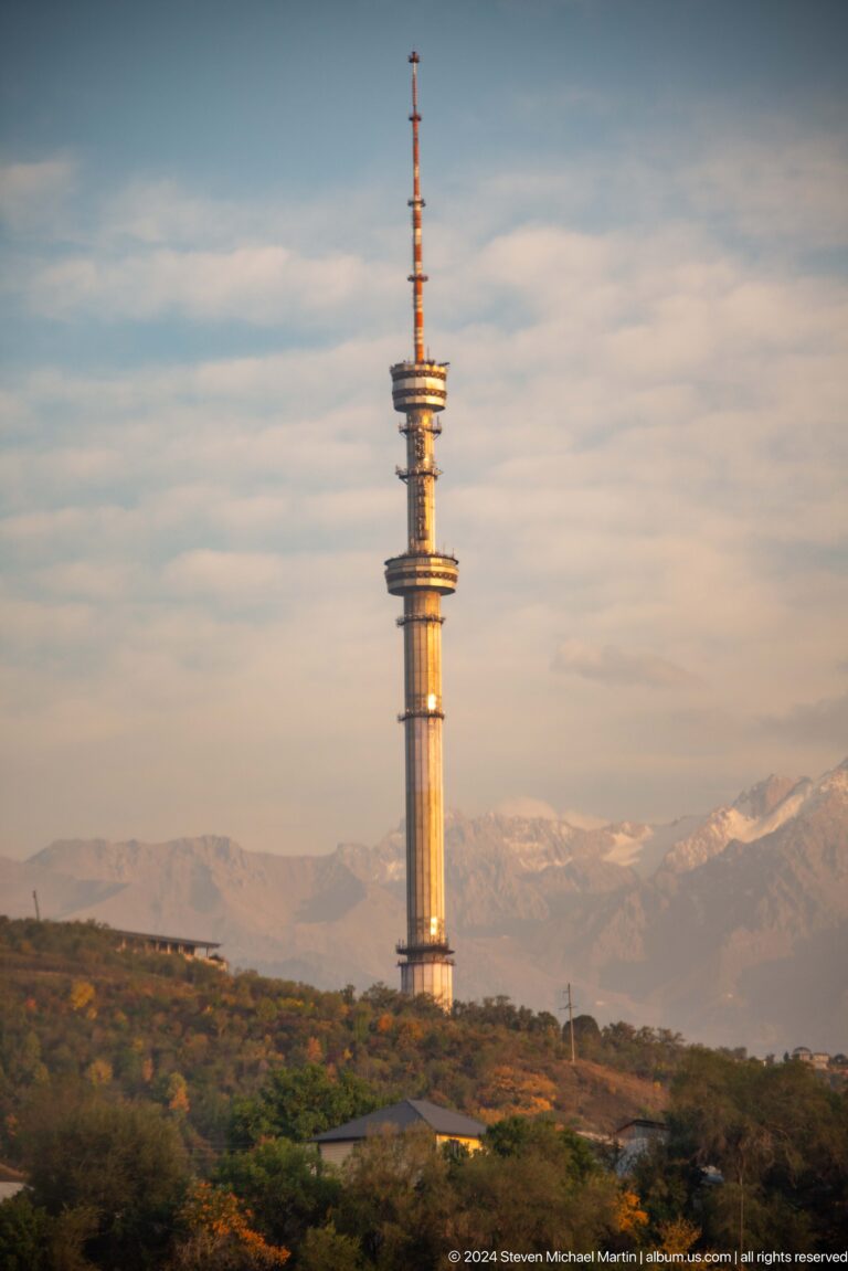 Upper section of the Almaty Television Tower with ringed platforms standing before a distant snow-covered Tian Shan mountain ridge.