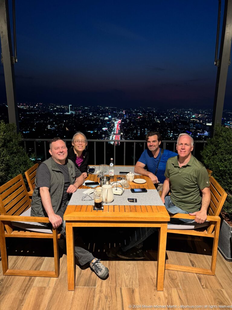 Four visitors sit at an outdoor Kok Tobe restaurant table at night with the illuminated Almaty city skyline in the background.