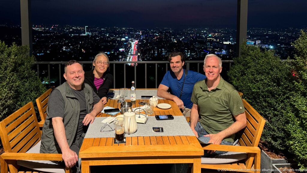 Four adults smiling around a wooden table on an elevated Kok Tobe restaurant terrace at night with Almaty city lights in the background.