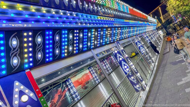 Nighttime view of a long LED-lit amusement attraction facade at Kok Tobe with multicolored lights and reflective panels.
