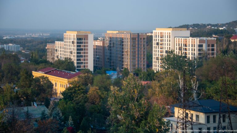 Elevated view from Kok Tobe of modern residential buildings and a yellow historic-looking structure surrounded by dense trees in Almaty.