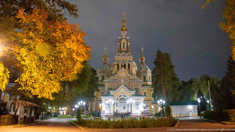 Night exterior framed by autumn trees at the colorful wooden Zenkov Cathedral in Almaty.