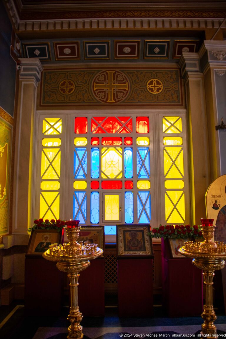 Stained-glass window above candle stands inside Zenkov Cathedral, with Orthodox religious decoration.