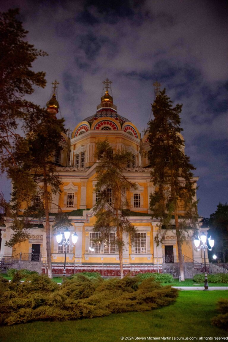 Night side view partly screened by trees at the colorful wooden Zenkov Cathedral in Almaty.