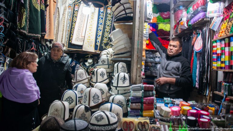 Market vendor surrounded by hats and textiles in Bishkek, Kyrgyzstan.