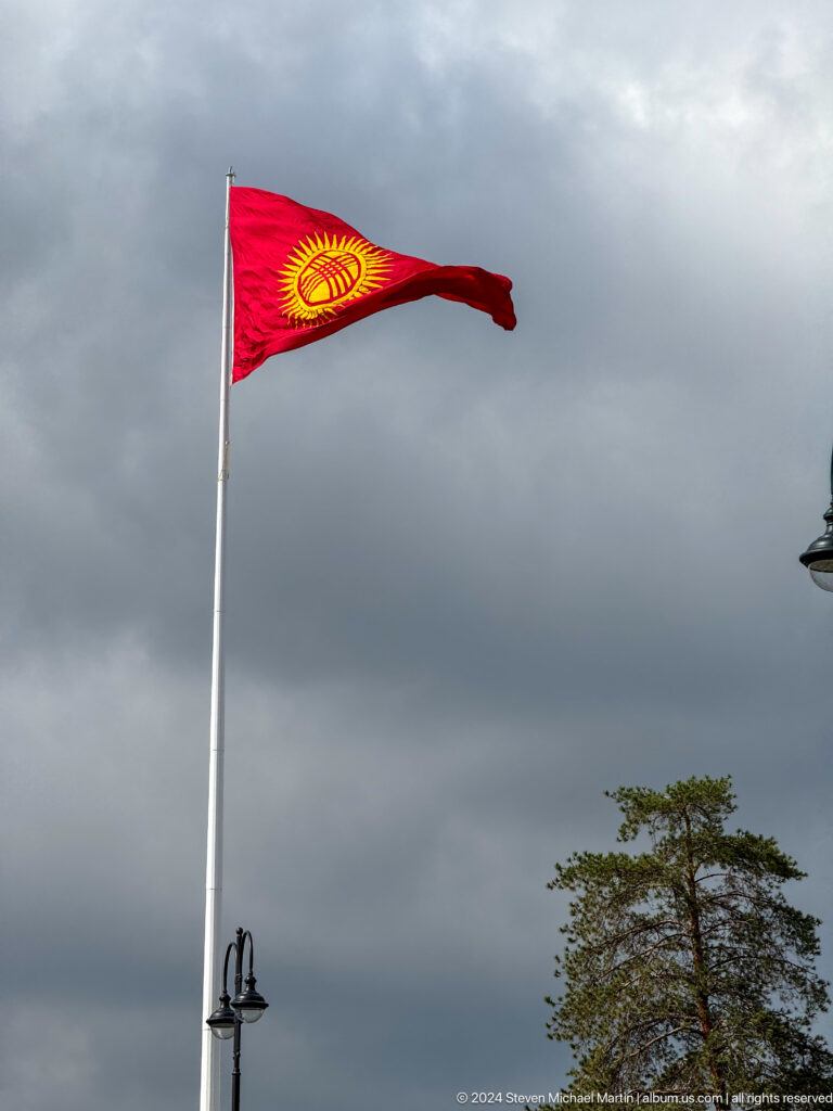 Kyrgyz national flag flying from a tall pole beneath storm clouds.