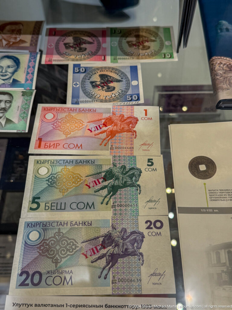 Small-denomination Kyrgyz banknotes beside a dark coin or disk.