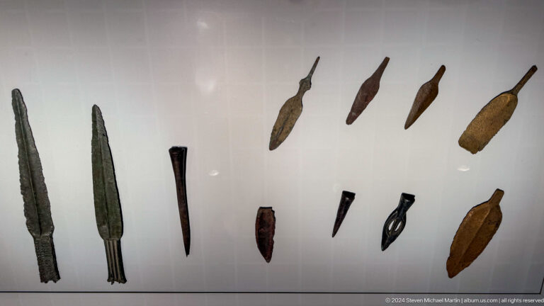 Row of bronze arrowheads and small blades on a wall mount.